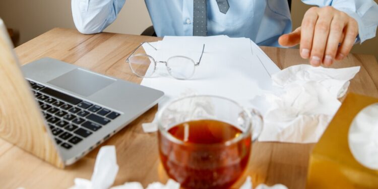 Feeling sick and tired. The man with cup of hot tea working in office, businessman caught cold, seasonal flu. Pandemic influenza, disease prevention, air conditioning in office cause sickness