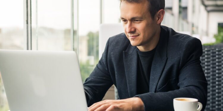 Using advantages of free Wi-Fi. Handsome young man working on laptop and smiling while sitting in sidewalk cafe