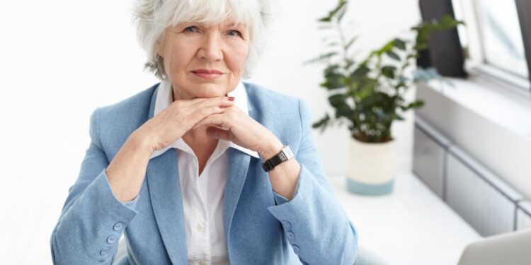 Horizontal shot of stylish elderly female real estate manager wearing nice blue suit and spectacles on her head, clasping hands under chin, having serious confident look, using laptop for work
