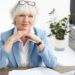 Horizontal shot of stylish elderly female real estate manager wearing nice blue suit and spectacles on her head, clasping hands under chin, having serious confident look, using laptop for work