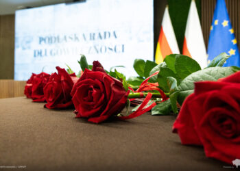 Czerwone róże na stole, w tle ekran z napisem Podlaska Rada Długowieczności oraz flagi Polski i Unii Europejskiej
