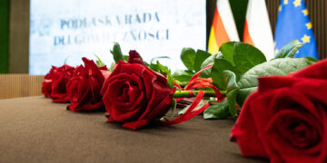 Czerwone róże na stole, w tle ekran z napisem Podlaska Rada Długowieczności oraz flagi Polski i Unii Europejskiej