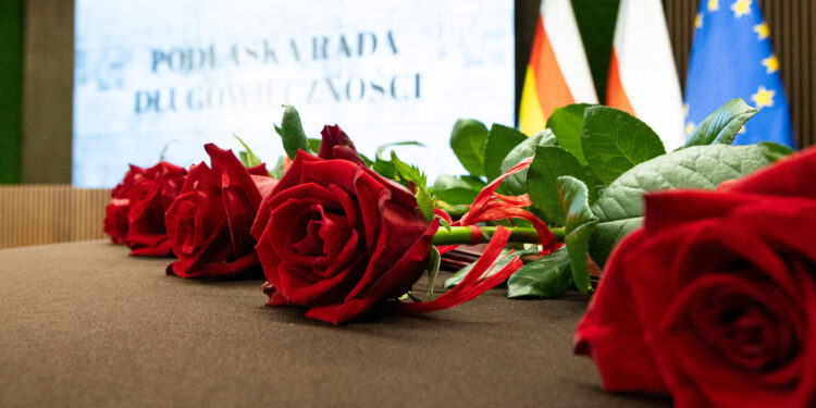 Czerwone róże na stole, w tle ekran z napisem Podlaska Rada Długowieczności oraz flagi Polski i Unii Europejskiej