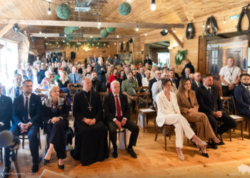 Goście jubileuszu dziesięciolecia Podlaskiego Muzeum Kultury Ludowej w drewnianej sali, w tle szyld muzeum