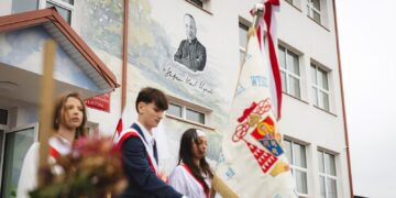 Poczet sztandarowy przed budynkiem szkoły podstawowej w Korycinie z muralem patrona