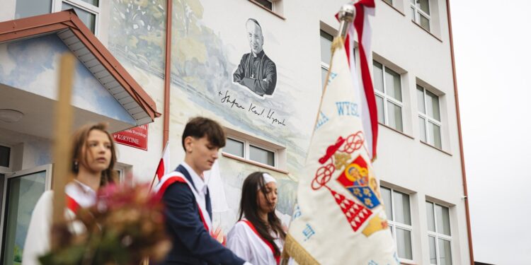 Poczet sztandarowy przed budynkiem szkoły podstawowej w Korycinie z muralem patrona