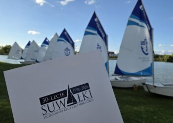 Optymisty z podniesionymi żaglami na brzegu Zalewu Arkadia w Suwałkach oraz tablica z napisem 30-lecie Uczniowski Klub Żeglarski Grot 1996-2026