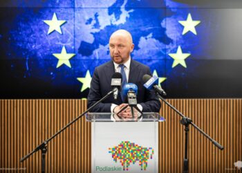 Marszałek województwa podlaskiego Łukasz Prokorym podczas konferencji prasowej na tle flagi Unii Europejskiej