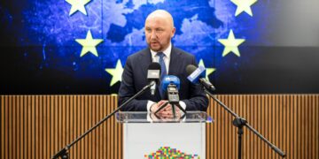 Marszałek województwa podlaskiego Łukasz Prokorym podczas konferencji prasowej na tle flagi Unii Europejskiej